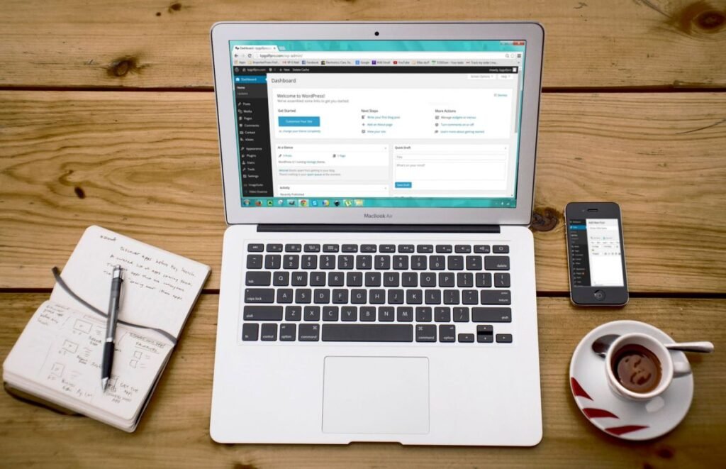 Portátil con el escritorio de WordPress abierto, junto a una libreta, un móvil y un café sobre una mesa de madera