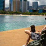 Partner Digital trabajando con un portátil en un banco frente al mar, con rascacielos al fondo