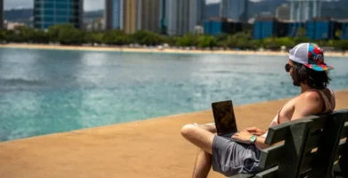 Partner Digital trabajando con un portátil en un banco frente al mar, con rascacielos al fondo